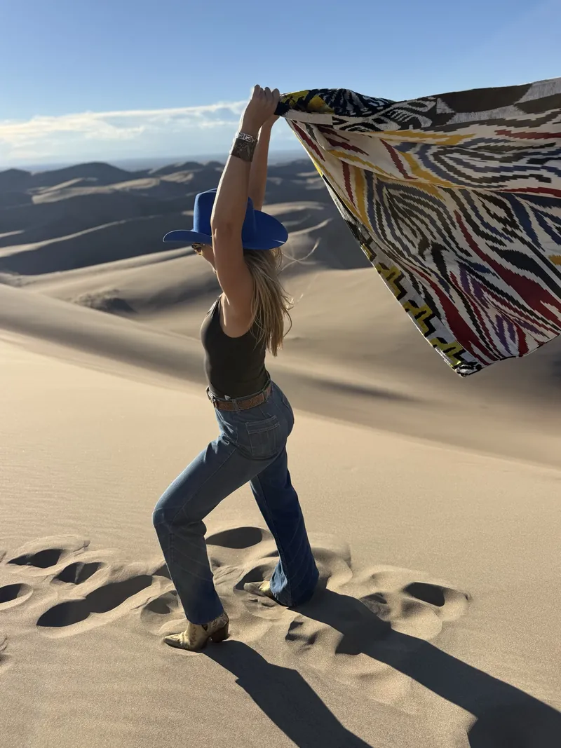A woman holds a patterned coat open like a sail against the wind on a sand dune.