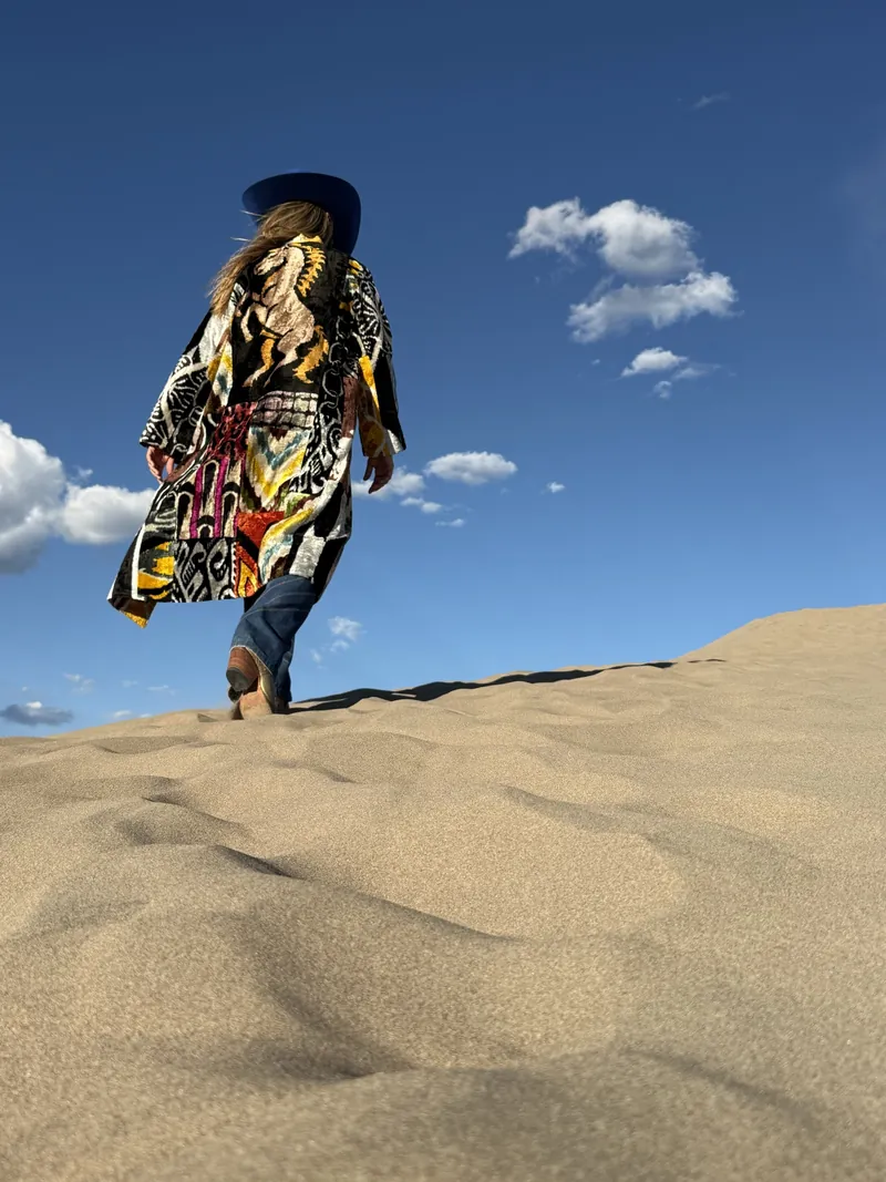 A woman in a patterned caftan walks up a sand dune toward a deep blue sky.