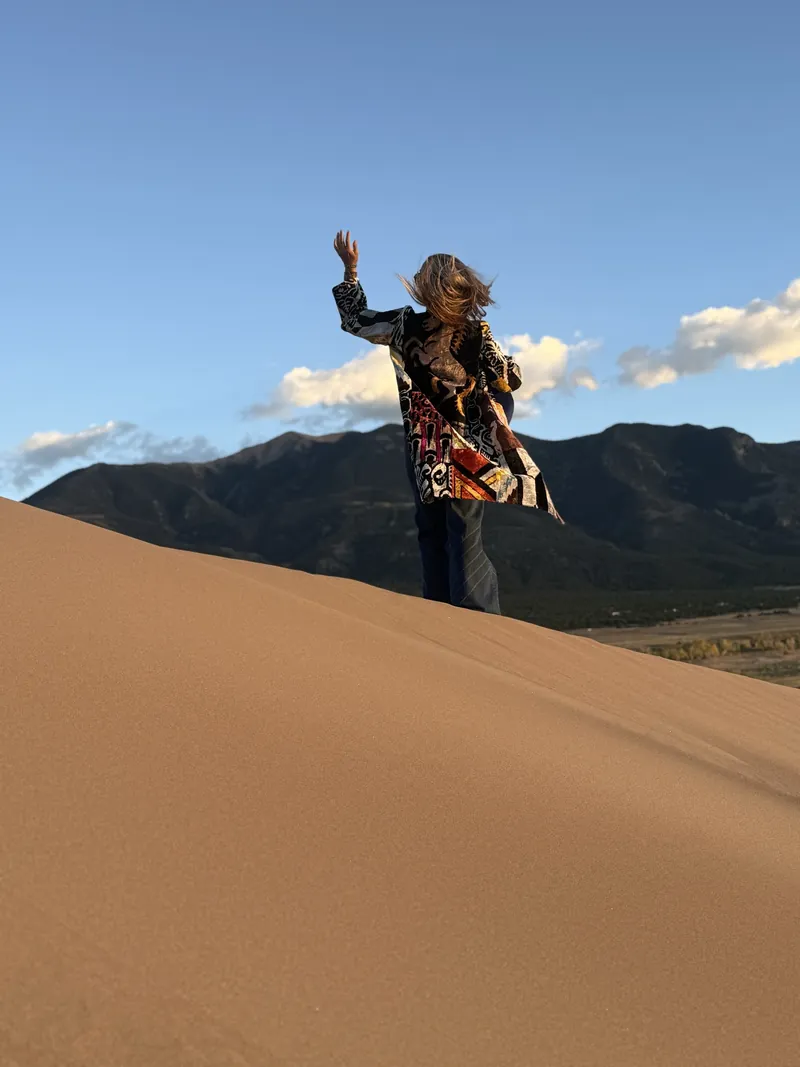 A woman with one arm raised stands on a dune crest in a patterned caftan, with mountains and a blue sky behind her.
