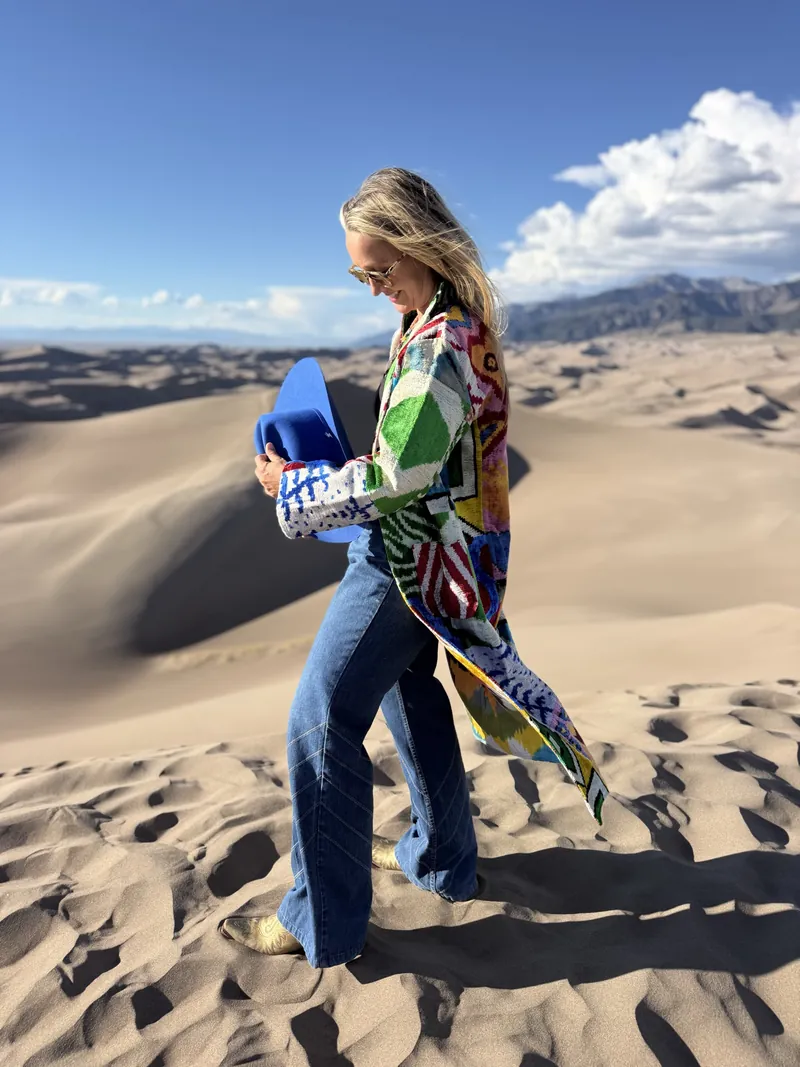 A woman in a patterned caftan walks across sand holding her cobalt hat against her side.