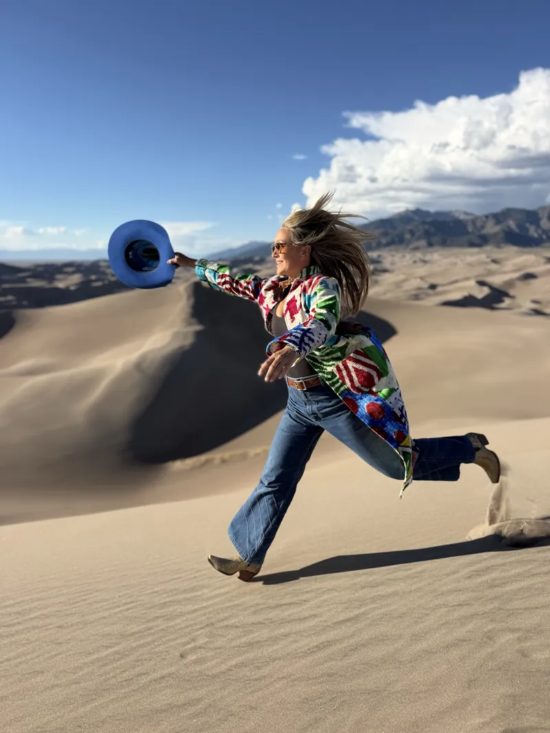 A woman in a multicolored ikat coat runs across a sand dune, tossing her cobalt cowboy hat into the air.