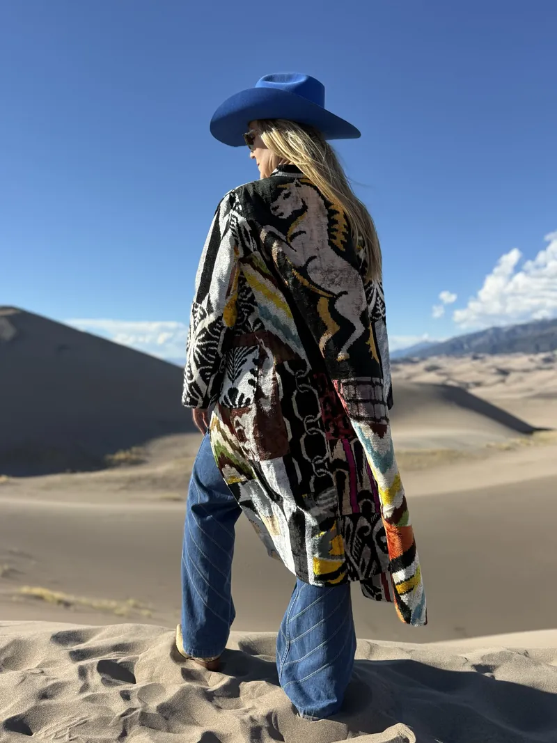 A woman in a cobalt hat and a black, white and red patchwork coat turns her head to the right, standing on a dune ridge under a deep blue sky.