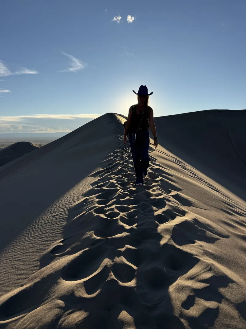A lone figure walks a dune ridge at low sun, casting a long shadow across rippled sand.