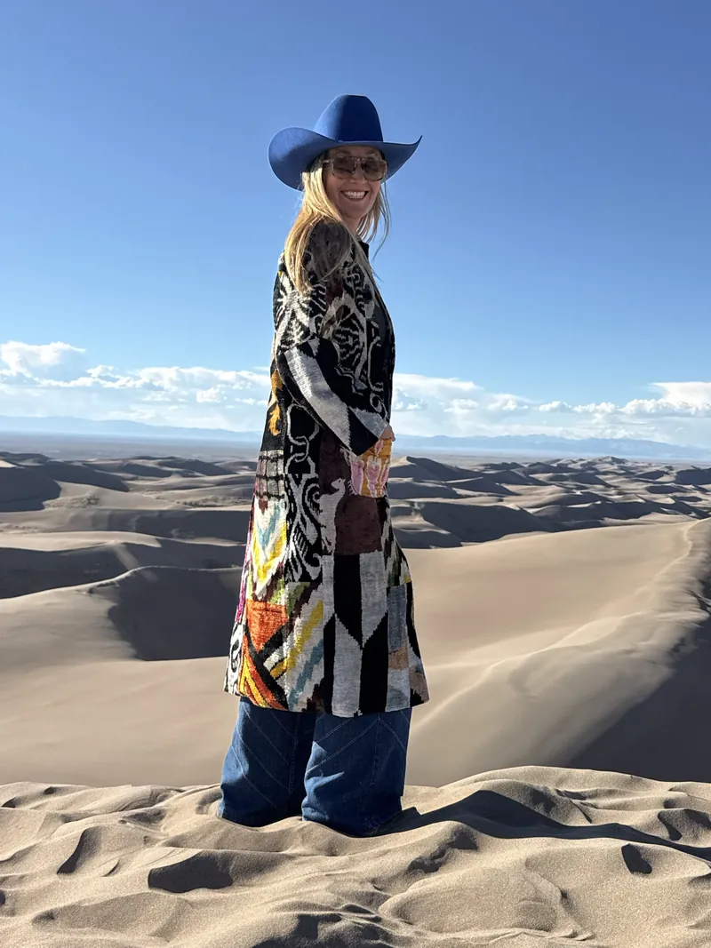 A smiling woman in sunglasses, a cobalt hat and a long black-and-white patterned caftan stands on a sand dune, with more dunes and distant mountains behind her.