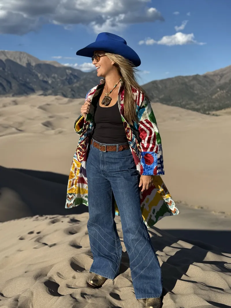 A smiling woman in a cobalt hat and a colorful ikat coat, hands on the lapels, stands on a dune with mountains behind her.