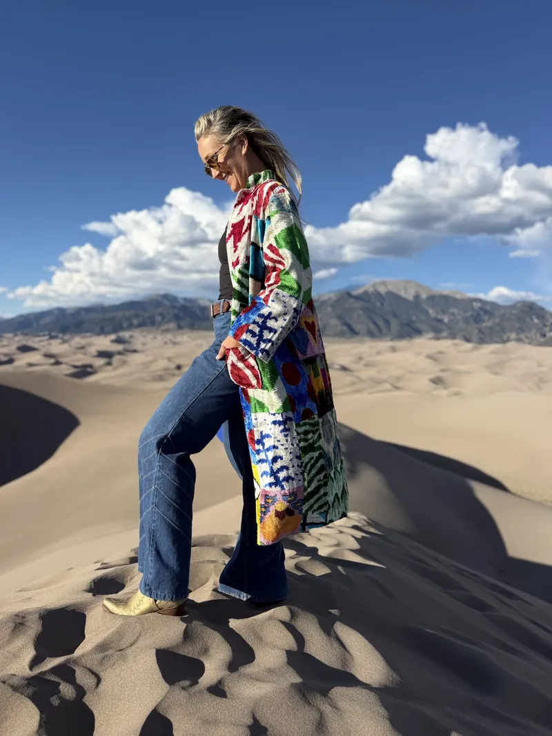 A woman in a red, green and blue patterned caftan steps along a dune ridge, looking down.
