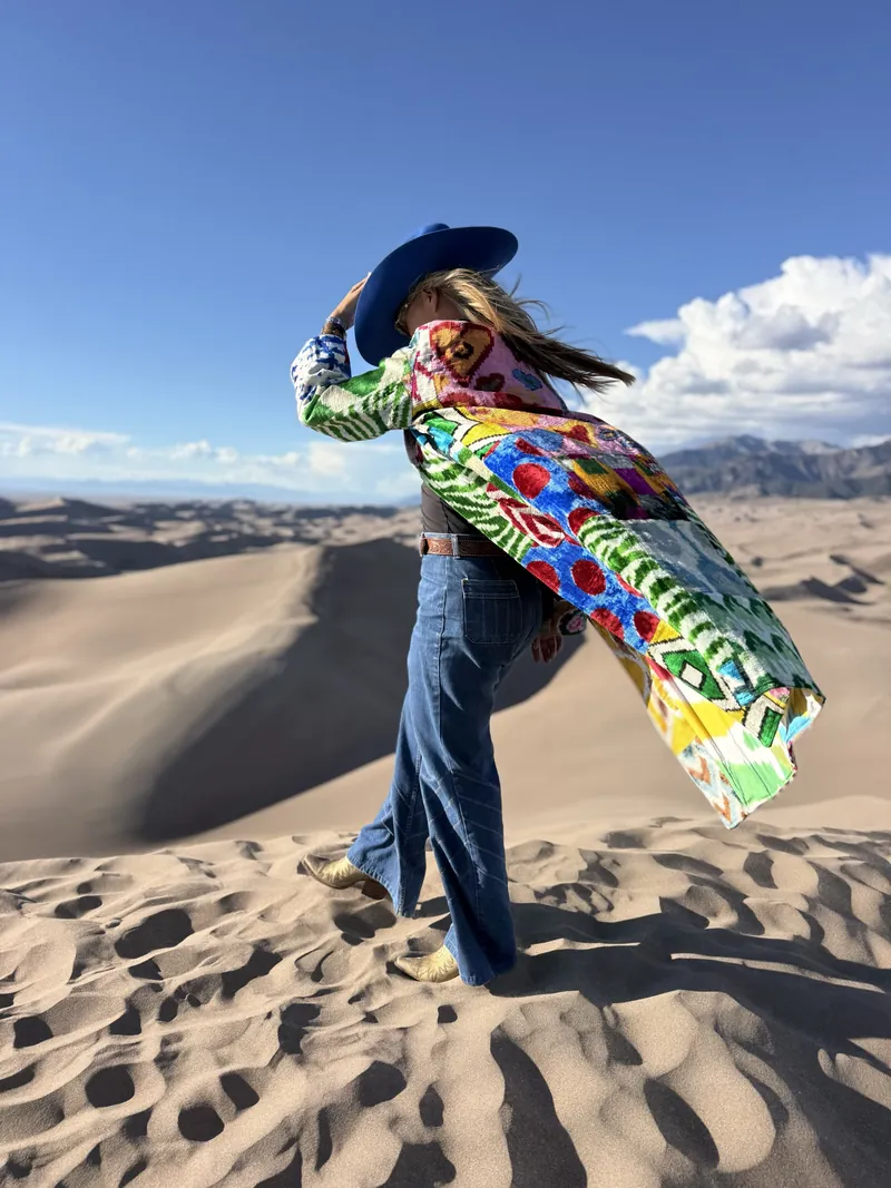 A woman in a brightly patterned caftan leans into the wind on a sand dune, one hand holding her cobalt hat.
