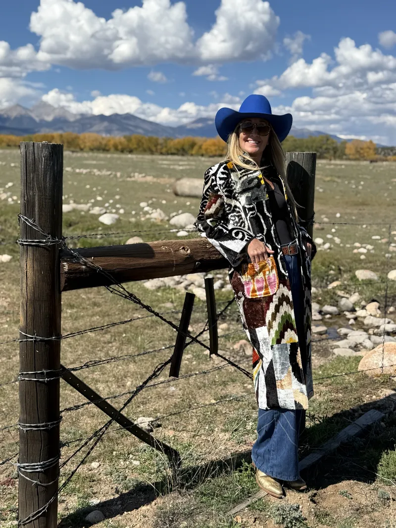 A woman in a cobalt hat and a black-and-white patterned caftan leans against a wooden fence post in a rocky pasture, with autumn trees and mountains behind.