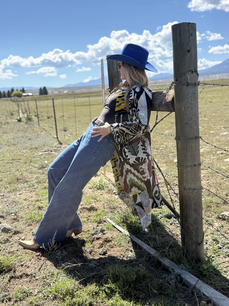 A woman in a cobalt hat leans back against a fence post, one leg crossed, in a green pasture.
