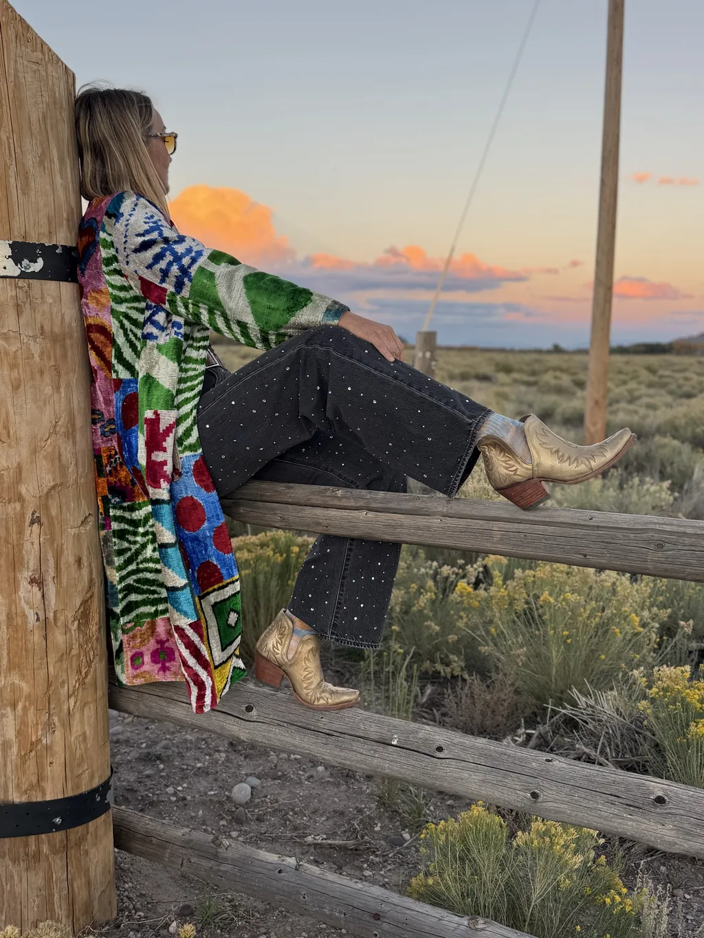 At sunset, a woman in a bright patchwork coat and gold boots leans against a fence post, looking out across a sagebrush plain.