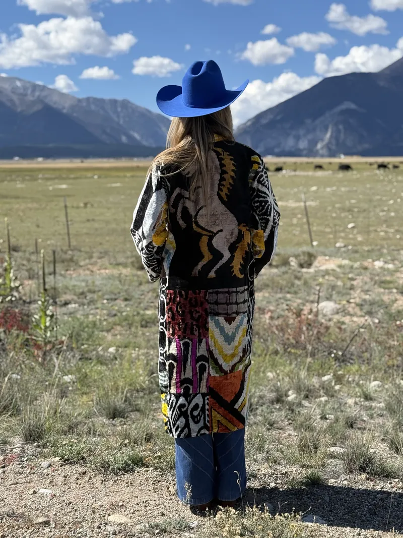Seen from behind, a woman in a cobalt hat and a horse-motif patterned caftan stands in a meadow with mountains and a blue sky ahead.