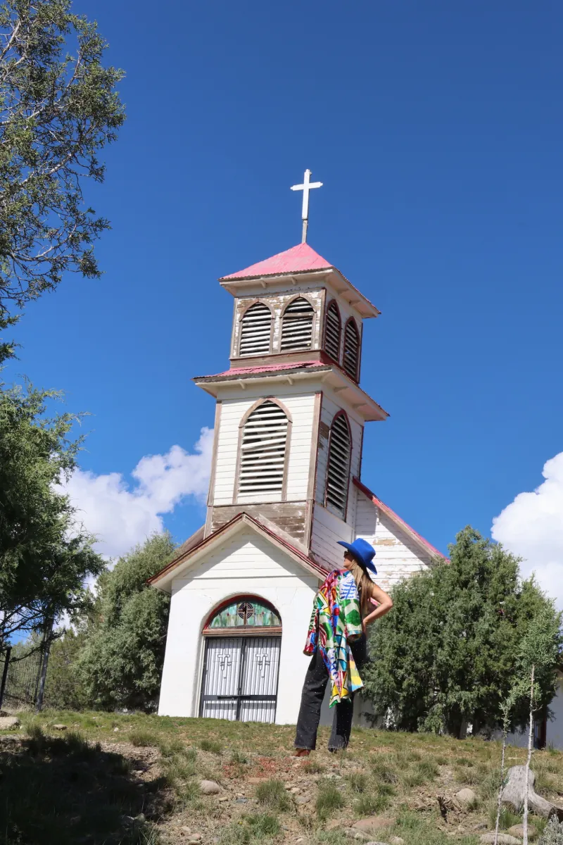 A woman in a patchwork caftan stands before the weathered bell tower and cross of St. John the Baptist Mission Church under a clear blue sky.