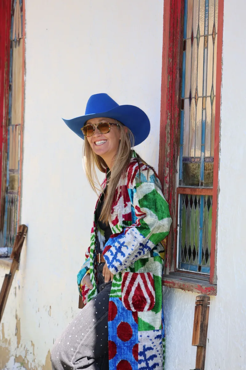 Smiling, a woman in a cobalt hat and patchwork caftan leans against the mission's whitewashed wall beside a red-trimmed stained glass window.