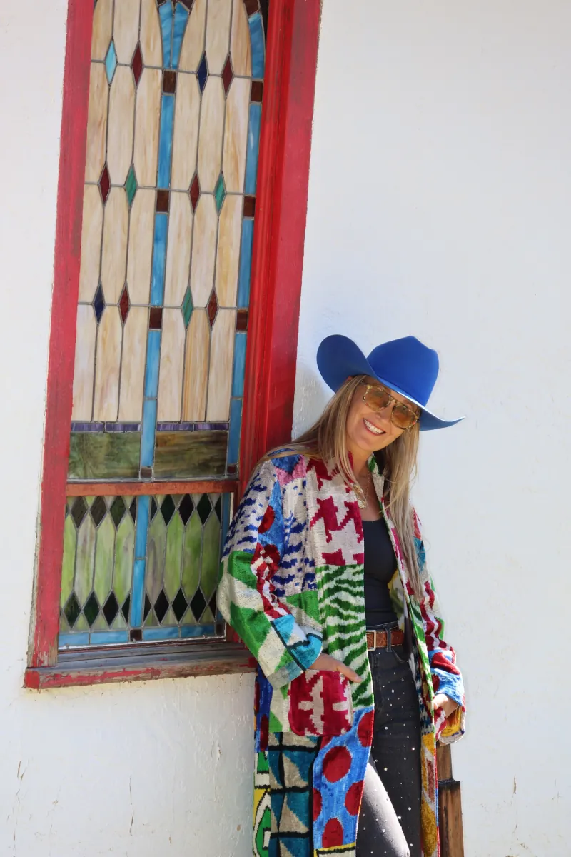 A woman in a cobalt hat and patchwork caftan smiles beside a tall stained glass window framed in weathered red paint at the mission.