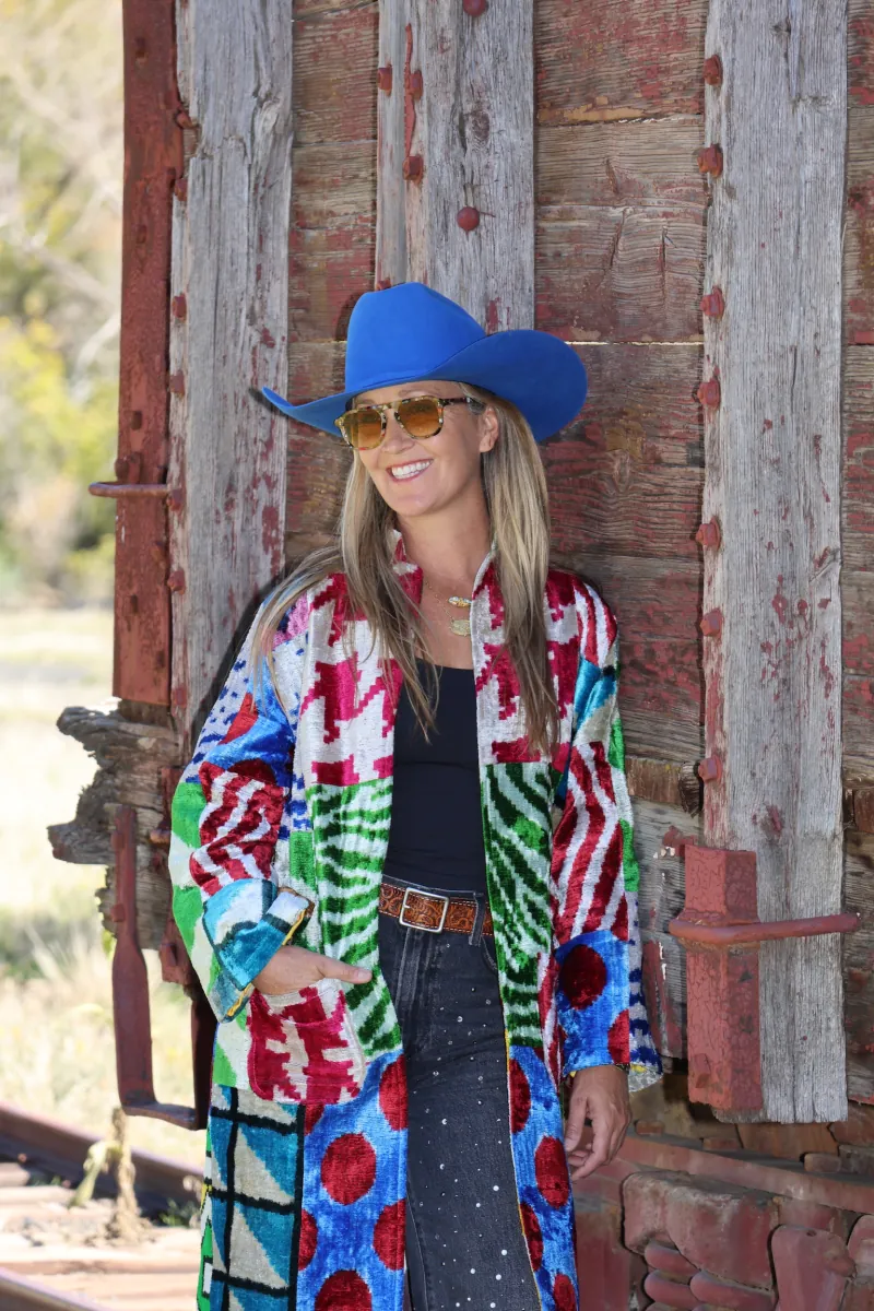 A woman in a patchwork caftan and cobalt hat leans against a weathered, rust-red railcar, hand in her pocket.