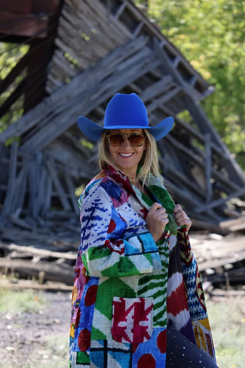 Smiling, a woman in a cobalt cowboy hat holds open her patchwork caftan in front of a collapsed wooden railroad structure.