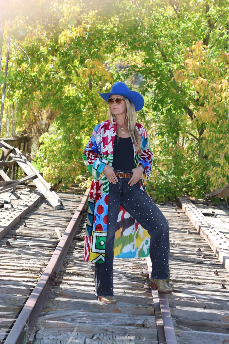 A woman in a cobalt cowboy hat and a colorful patchwork caftan stands on a weathered railroad trestle, autumn trees behind her.