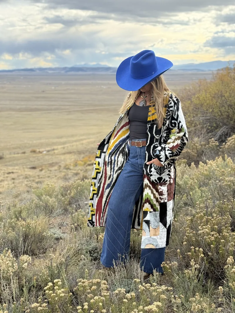 A woman in a cobalt hat and a black, white and red patterned caftan stands among golden shrubs on a wide plain, hands in her pockets.