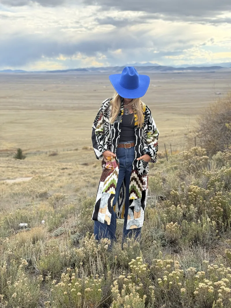 A woman in a cobalt hat tipped low and an ikat coat stands in a field of golden shrubs.