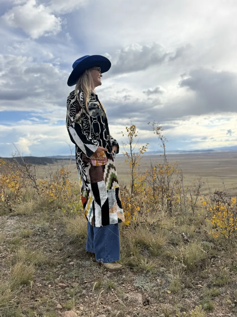 A woman in a cobalt hat and a black and white coat looks out over an autumn prairie under a broad cloudy sky.